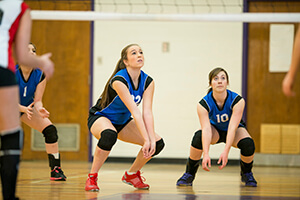 Two female volleyball players crouching below a net waiting for the ball.