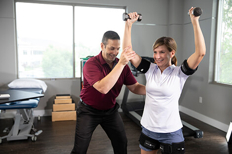 Male therapist spotting a female patient raising small dumbbells over her head.