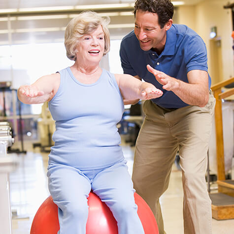 Male therapist supervising woman doing balance exercise on large orange ball