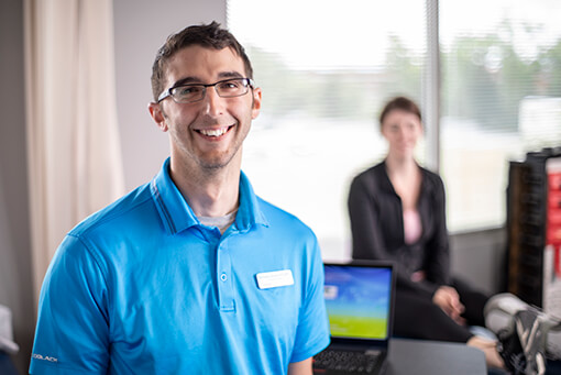 therapist wearing a teal blue shirt and name tag standing in front of a laptop on a desk