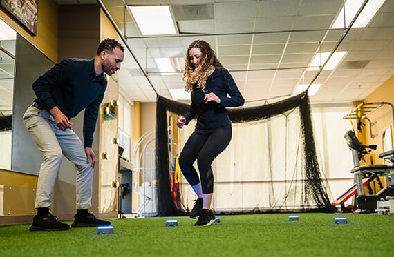 An athlete is performing a reaction drill with light up buttons on the ground while a therapist is standing beside her