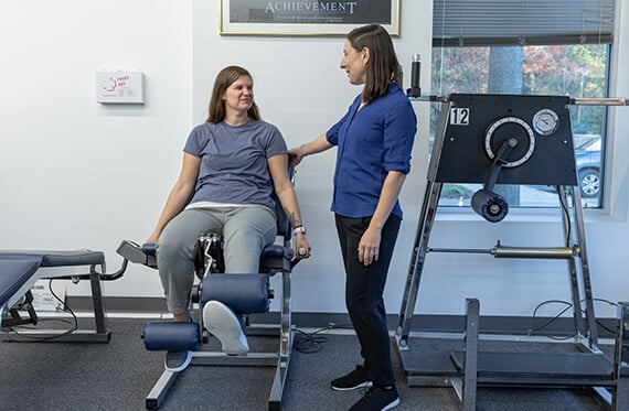 A woman is raising one leg on a leg extension machine while a therapist is standing beside  for support