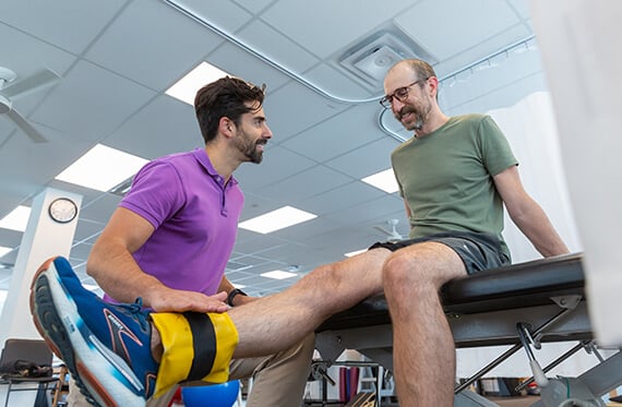 A man is sitting on a bench and raising one leg that has a weight around the ankle. A therapist is kneeling beside and smiling at him